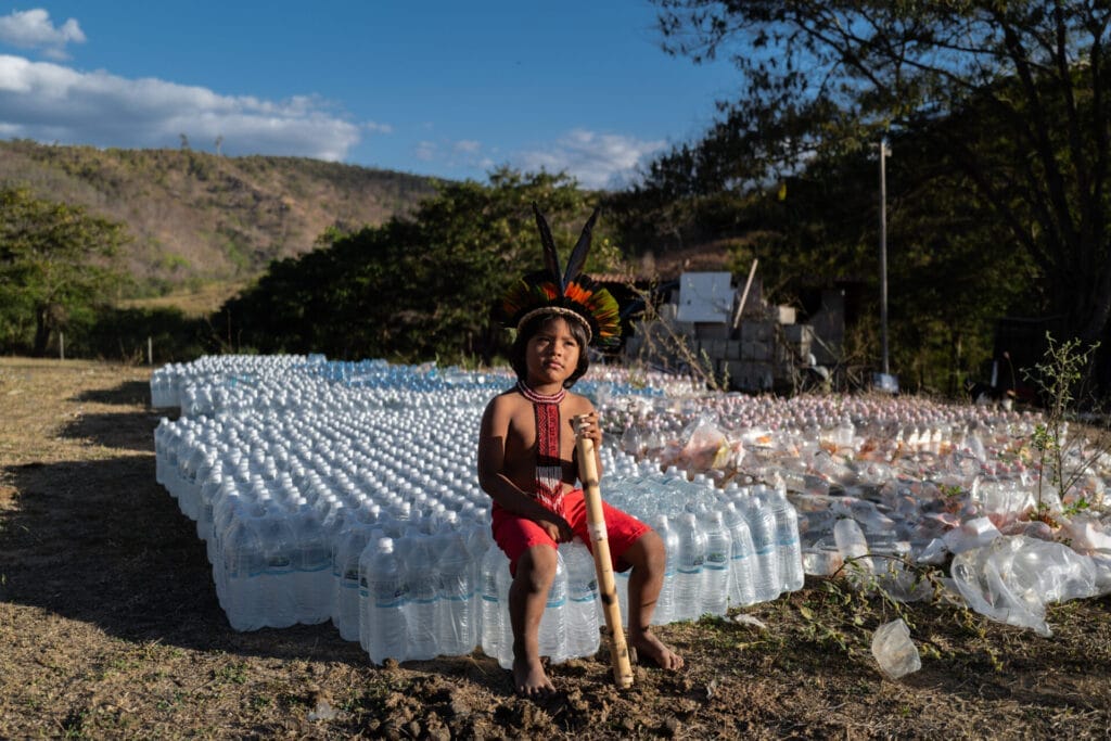 Dhombre Krenak, com roupas tradicionais, sentado sobre o estoque de garrafas de água potável no quintal da escola estadual indígena na terra indígena Krenak, no estado de Minas Gerais.